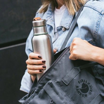 Stainless Steel drinkfles van Black&Blum met dop met groen rand en leder draagbandje die in een jeans tas gestoken wordt door dame met bruine nagellak